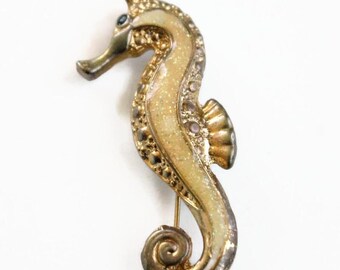 May include: A seahorse-shaped brooch featuring a metallic gold-tone finish with a textured, glittery cream-colored inlay along the body. The design includes small, embedded clear stones and a dark, circular eye detail on the head of the seahorse.