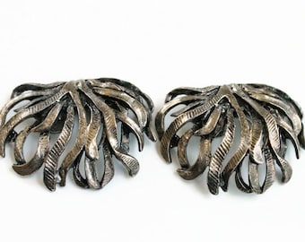 May include: A pair of silver-toned metal shoe clips featuring a textured, fan-like design with multiple curved, ribbon-shaped segments radiating from a central point. The metallic surface has a dark, antiqued finish with visible etched lines.
