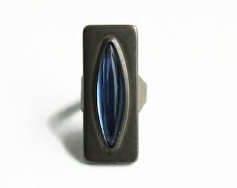 May include: A rectangular ring featuring a dark, matte metallic finish and a central, elongated marquise-shaped blue stone. The stone is set vertically within the center of the metal plate, which has rounded corners and a smooth, flat surface.