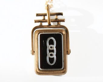 May include: A vintage gold-toned pendant necklace featuring a rectangular black enamel center with a silver-colored interlocking rings design. The pendant is suspended from a delicate gold chain. The necklace has a classic, retro aesthetic.