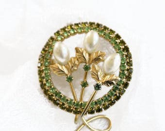 May include: A circular brooch featuring a border of small green rhinestones surrounding three gold-toned stems. Each stem is topped with a white faux pearl and gold-toned leaves, with additional green rhinestones accenting the stems and leaves.