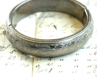 May include: A silver-toned metal hinged bangle bracelet featuring an intricate engraved floral pattern across its outer surface. The circular bracelet has a visible hinge mechanism on the left side and a smooth interior finish with a metallic sheen.