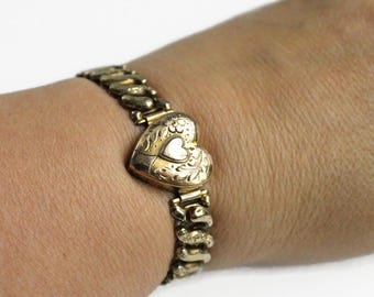 May include: A gold-toned metal link bracelet featuring a central heart-shaped locket. The locket is decorated with an engraved floral pattern and a smaller, smooth heart shape in the center. The bracelet consists of curved, interlocking metal links.