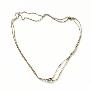 May include: A delicate, gold-colored necklace with two strands of fine chain. The necklace is laid out on a white surface, with a clasp visible at the bottom. The chains are connected at the clasp.