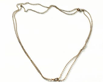May include: A delicate, gold-colored necklace with two strands of fine chain. The necklace is laid out on a white surface, with a clasp visible at the bottom. The chains are connected at the clasp.