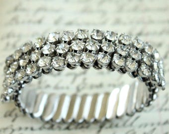 May include: A silver-toned stretch bracelet featuring three parallel rows of clear, round-cut rhinestones set in metal prongs. The band is constructed with a flexible, expandable metal link design that allows it to stretch for a comfortable fit.