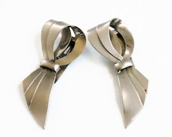 May include: A pair of silver-toned metal earrings shaped like stylized bows. Each earring features a smooth, curved loop at the top that transitions into a flat, tapered tail section with subtle linear detailing, creating a polished metallic finish.
