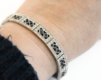 May include: Silver-toned bracelet with a geometric design. Each rectangular link features a black diamond-shaped center, surrounded by clear, sparkling crystals. The bracelet is worn on a wrist, with a dark blue sleeve visible.