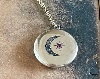 May include: A gold chain necklace with a round locket pendant featuring a crescent moon design with black stones and a red gemstone in the center.