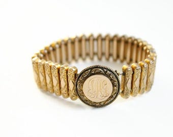 May include: A gold-toned bracelet with an engraved central medallion. The bracelet features a series of rectangular links with decorative patterns. The medallion displays an engraved monogram. The bracelet is set against a white background.