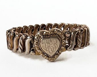 May include: A metallic expansion bracelet featuring a central heart-shaped charm with an ornate border and engraved script initials. The band consists of repeating S-shaped links with a textured finish, creating a flexible and continuous circular design.