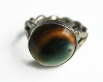 May include: A silver-toned ring featuring a round, smooth cabochon stone with a gradient pattern transitioning from dark brown to a deep black center and a soft green base. The metal band has a twisted, rope-like texture surrounding the stone.
