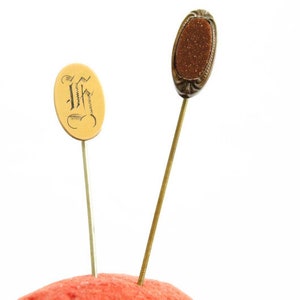 May include: Two decorative pins with oval-shaped heads. One pin has a gold head with black lettering, the other has a brown head with a gold rim.