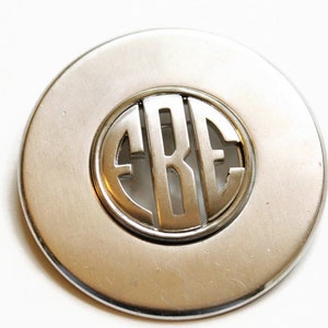May include: A silver circular brooch with a monogram of the letters "EBE" cut out of the centre.