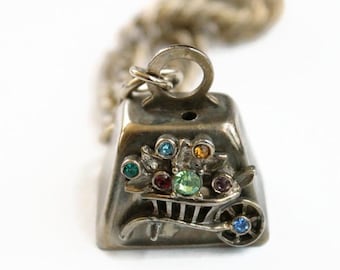 May include: A silver-toned metal charm in the shape of a wheelbarrow, featuring a top loop for attachment and a chain. The wheelbarrow is decorated with small, multi-colored inset rhinestones in shades of green, blue, red, yellow, and purple.