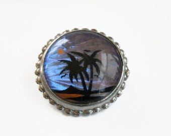 May include: A round brooch featuring a circular inlay of iridescent blue butterfly wing material. The center displays a black silhouette of two palm trees against a sunset sky with a small orange sun, all encased in a scalloped silver-tone metal frame.