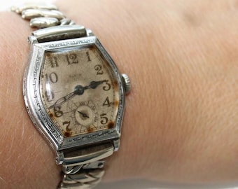 May include: A vintage silver-tone wristwatch with a hexagonal case and a metallic link bracelet. The off-white dial features black Arabic numerals, a small sub-dial for seconds at the bottom, and dark hands. The bezel has an engraved Greek key pattern.