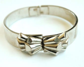 May include: A silver-toned metal hinged bangle bracelet featuring a central decorative element composed of two symmetrical, fan-shaped geometric clusters with raised, angular facets that meet at the center of the band.