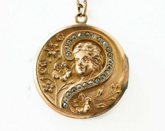 Pode incluir: Um pingente de medalhão de ouro com um design floral detalhado e um rosto de mulher no centro. O medalhão é cercado por uma fileira de pequenas pedras transparentes.