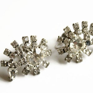 May include: A pair of silver clip-on earrings with a snowflake design. Each earring features a large, clear rhinestone surrounded by smaller rhinestones.