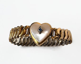 May include: A gold-toned expandable bracelet featuring a central heart-shaped mother-of-pearl plaque. The plaque is accented with a small, dark-set diamond-shaped element containing a single blue stone. The band consists of interlocking metal links.