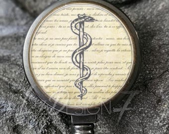 Könnte beinhalten: Schwarze Ausweisrolle mit bronzefarbenem Rahmen. Die Rolle hat ein kreisförmiges Design mit einem medizinischen Symbol und Schrift auf cremefarbenem Hintergrund. Die Rolle hat einen transparenten Kunststoffriemen und einen Metallclip.