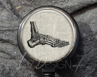 May include: A black and silver retractable badge holder with a round, silver button. The button has a black and white image of a human foot skeleton.