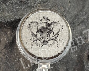 May include: A silver retractable badge holder with a round, clear plastic case featuring a black and white anatomical illustration of a human pelvis on a linen-textured background.