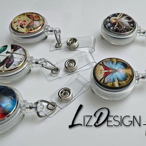 May include: Five retractable badge holders with different designs. Each holder features a round, clear plastic case with a colorful image inside. The images include a butterfly, a dragonfly, an octopus, a galaxy, and a fairy with butterfly wings. The holders are attached to clear plastic retractable cords with a metal clip.