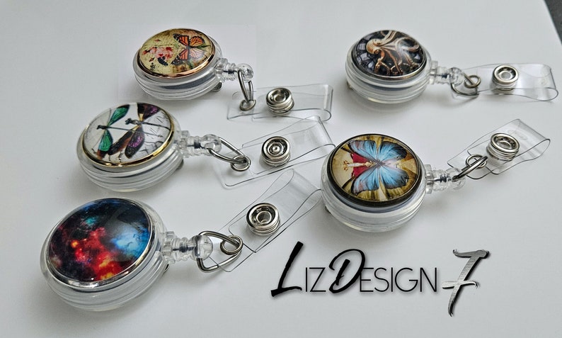 May include: Five retractable badge holders with different designs. Each holder features a round, clear plastic case with a colorful image inside. The images include a butterfly, a dragonfly, an octopus, a galaxy, and a fairy with butterfly wings. The holders are attached to clear plastic retractable cords with a metal clip.