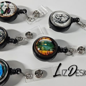 May include: Five different retractable badge holders with various designs. Each holder has a black plastic body with a clear plastic strap and a metal clip. The designs include a butterfly, an owl, a clock, a peacock feather, and a heart.