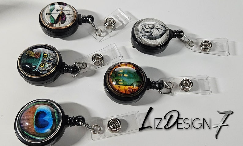 May include: Five different retractable badge holders with various designs. Each holder has a black plastic body with a clear plastic strap and a metal clip. The designs include a butterfly, an owl, a clock, a peacock feather, and a heart.