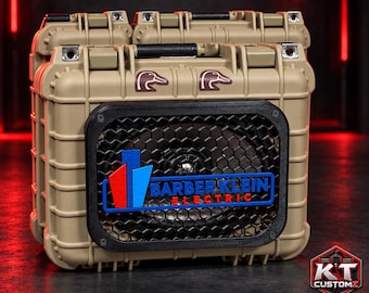 May include: A tan, rectangular, hard-shell protective case converted into a portable speaker. It features a black hexagonal grille, two brown duck head logos, and a blue and red logo reading BARBER KLEIN ELECTRIC on the front.