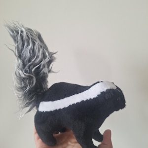 May include: A plush skunk toy featuring a soft black body with a white stripe running along the back and head. The skunk has a large, upright, fluffy tail with long, wavy grey and white synthetic fur fibres and small black bead eyes.