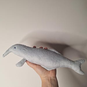 Pode incluir: Um brinquedo de pelúcia cinza claro em forma de golfinho, com um focinho longo, barbatanas pequenas e uma cauda. O brinquedo é segurado em uma mão contra um fundo simples. Os olhos do golfinho são pequenos e azuis.