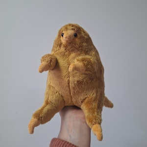 May include: A soft, plush toy resembling a creature with a rounded body, small arms, and legs. The toy is a warm, golden-brown colour, with a slightly lighter shade on its face and limbs. It has small, dark eyes and a soft, fuzzy texture.