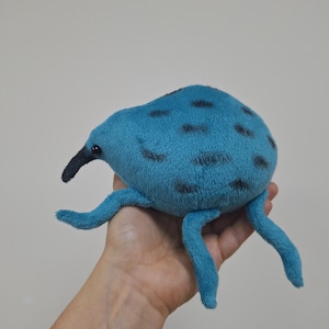 May include: A plush toy in the shape of a blue insect with black spots and four blue legs. The toy has a black beak and is held in a person's hand. The background is a plain, light gray color.