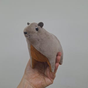 Puede incluir: Un juguete de peluche con forma de capibara. El juguete es marrón y gris con la barriga marrón. Tiene pequeños ojos y orejas negros.