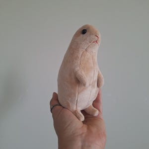May include: A small, light tan plush toy shaped like a standing rodent. The stuffed animal features a smooth, velvety texture, a rounded body, tiny front paws, and small hind legs. It has a simple face with two dark, round eyes and a stitched red mouth.
