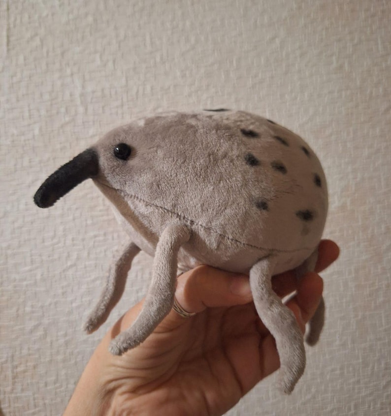 May include: A plush, gray insect toy with a long, black snout and small black spots. The stuffed animal has six gray legs and is held in a hand. The toy is made of soft fabric.