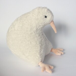 May include: A plush, off-white kiwi bird stuffed animal. The bird has a long, light pink beak and small, light pink feet. The soft, textured body is in a sitting position, with a small black eye visible.