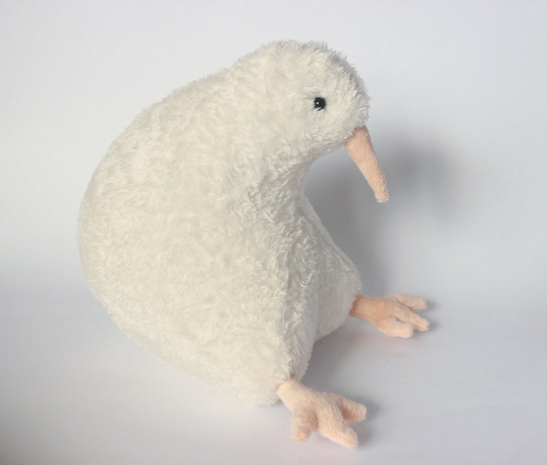May include: A plush, off-white kiwi bird stuffed animal. The bird has a long, light pink beak and small, light pink feet. The soft, textured body is in a sitting position, with a small black eye visible.
