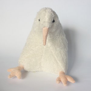 May include: A plush, white kiwi bird stuffed animal with a long, light pink beak and small, light pink feet. The soft toy has a fluffy texture and is sitting upright, with its feet extended.