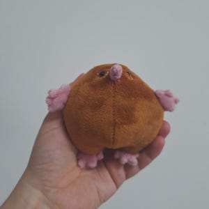 May include: A soft toy mole with a rounded brown body, small pink arms, feet, and nose. The stuffed animal is held in a hand against a plain white background. The mole has small black eyes and a stitched design.