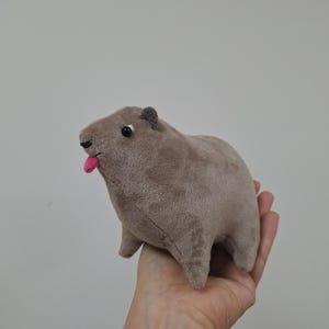 Puede incluir: Un juguete de peluche capibara marrón con la lengua rosa que sobresale. El juguete tiene ojos negros y orejas pequeñas.