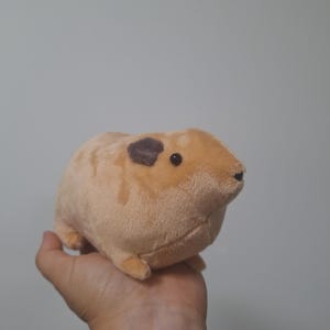 Puede incluir: Un juguete de peluche con forma de cobaya que presenta un pelaje suave de color tostado, un cuerpo redondeado, orejas pequeñas de color marrón oscuro y ojos de plástico negro. El peluche tiene una silueta compacta y robusta con extremidades.