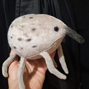 May include: A plush toy creature with a light gray body, black spots, and a long, dark gray snout. It has four gray legs and a black eye. The toy is soft and appears to be made of a plush material.