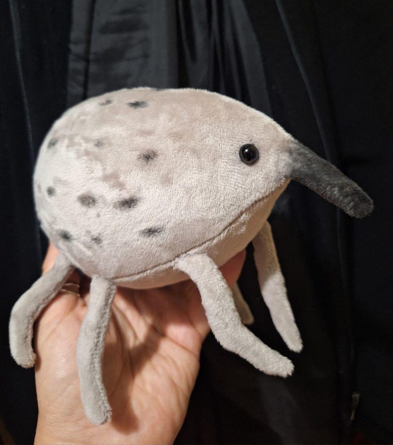May include: A plush toy creature with a light gray body, black spots, and a long, dark gray snout. It has four gray legs and a black eye. The toy is soft and appears to be made of a plush material.