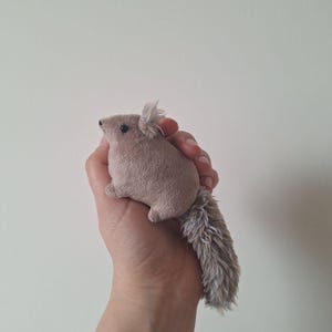 Op de afbeelding: Een klein, pluche bruin en grijs eekhoorn speelgoed met een lange, pluche staart. Het speelgoed wordt in de hand van een persoon gehouden.
