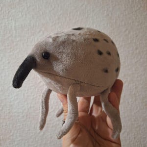 May include: A plush toy insect with a long black snout, gray body, and small black spots. The toy has three pairs of gray legs and small black eyes. The toy is made of soft, fuzzy material.